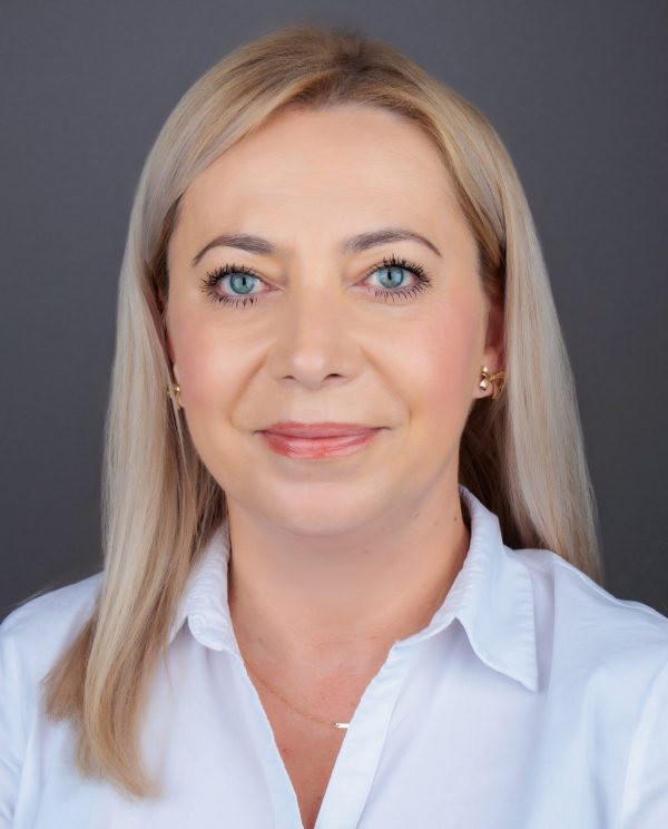 Smiling person with straight blonde hair and blue eyes wearing a white collared shirt, gold earrings, and a delicate necklace against a plain gray background.