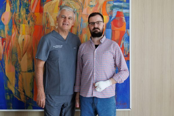 Dvojica muškaraca stoje ispred velikog, šarenog apstraktnog platna; lijevi u sivim medicinskim odorama, a desni bradati s naočalama u kariranoj košulji i sa zavojem na desnoj ruci.