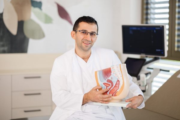 Liječnik u bijeloj kuti s naočalama sjedi u ordinaciji i drži anatomski model ženskog zdjeličnog područja, s monitorom ultrazvuka u pozadini.