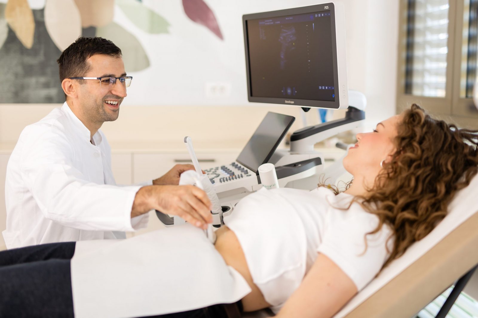 Liječnik u bijeloj kuti izvodi ultrazvučni pregled trbuha na pacijentici koja leži na stolu, dok se slika pregleda prikazuje na monitoru u svijetloj ambulanti.