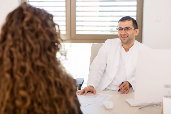 Liječnik u bijeloj kuti nasmiješeno razgovara s pacijenticom preko stola u ordinaciji, s papirima i računalom na stolu.