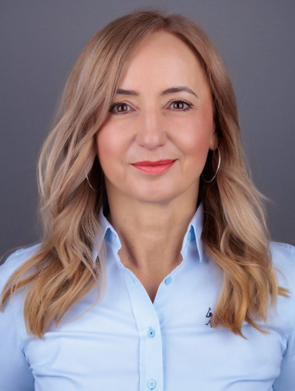 Portret žene s valovitom plavom kosom, okruglim naušnicama i blagim osmijehom, u svijetloplavoj košulji na sivoj pozadini.