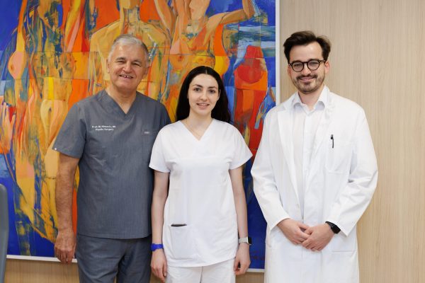 Troje medicinskih stručnjaka u uniformama i laboratorijskom kaputu, nasmiješeni, ispred šarene apstraktne slike.