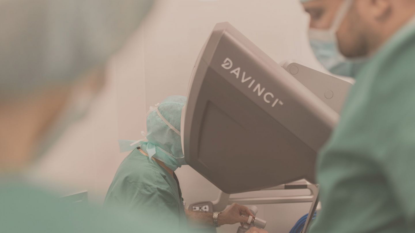 Medical professional at a da Vinci robotic surgical system console, with masked staff in green scrubs in the foreground.