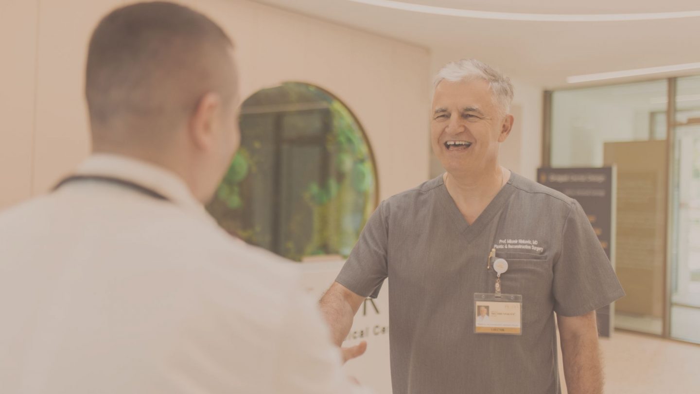 Smiling clinician in gray scrubs with ID badge shakes hands with a colleague in a white coat in a hospital hallway.