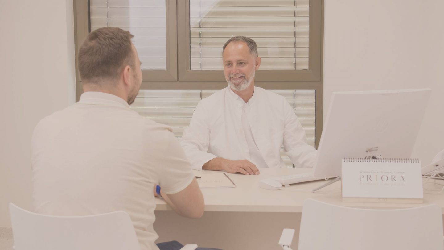 Doctor in a white coat smiles while consulting with a patient across a desk in a bright medical office with a computer.