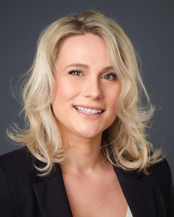 Portret nasmiješene osobe s valovitom plavom kosom u tamnom sakou na neutralnoj sivoj pozadini.