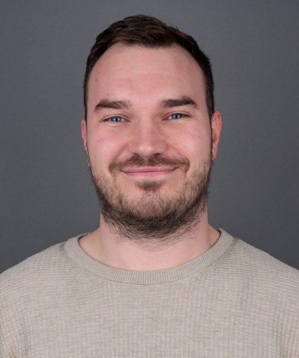 Osoba s kratkom smeđom kosom, plavim očima i kratkom bradom, u bež pletenoj majici, blago se smiješi pred sivom pozadinom.