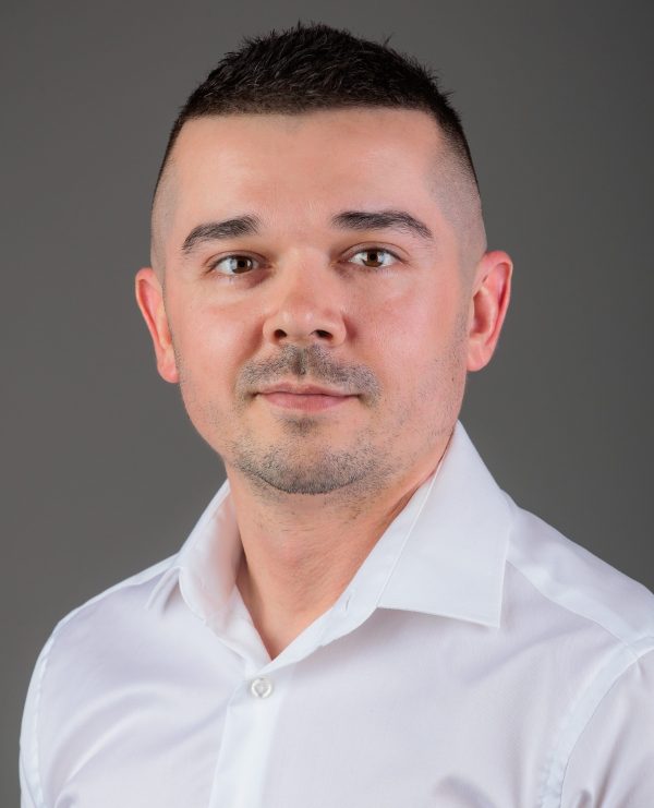 Portret muškarca kratke tamne kose i blage brade, u bijeloj košulji, na sivkastoj pozadini.