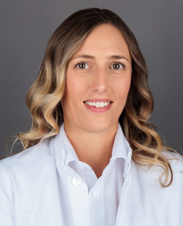 Žena u bijeloj medicinskoj kuti s valovitom svjetlosmeđom kosom, portret s blagim osmijehom na sivoj pozadini.