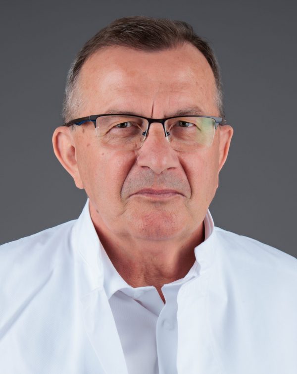 Stariji muškarac s naočalama u bijeloj kuti, ozbiljan izraz lica na sivoj pozadini.