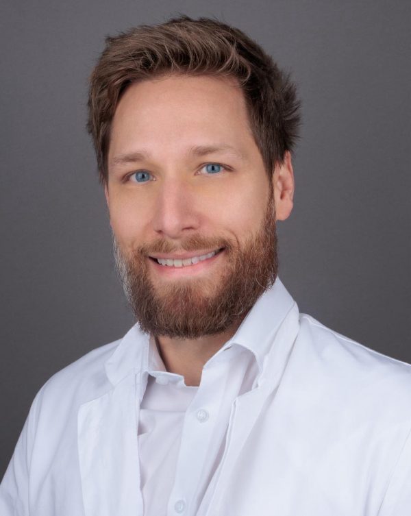 Portret muškarca s bradom i plavim očima u bijeloj liječničkoj kuti, nasmiješen ispred sive pozadine.