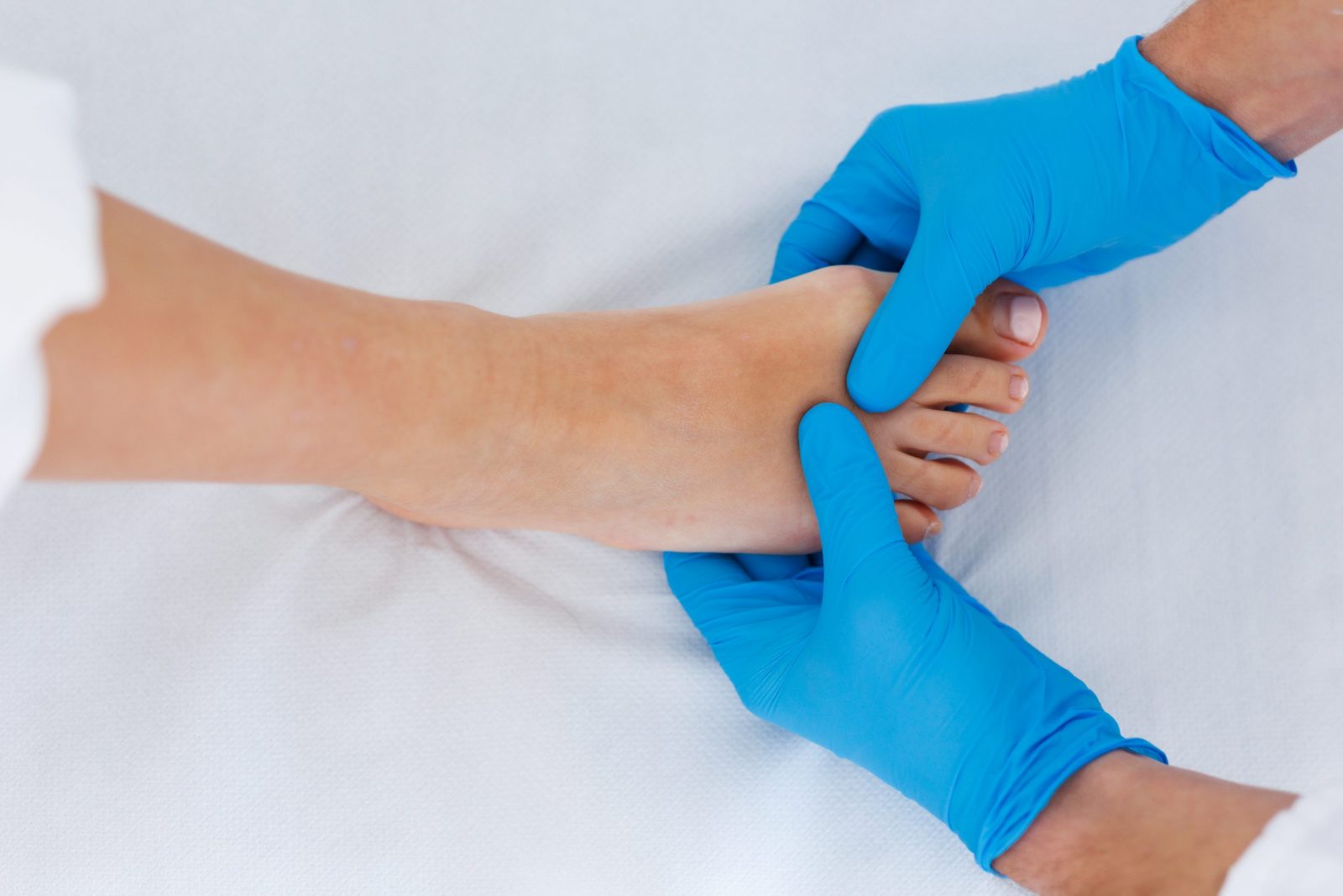 Stopalo koje pregledavaju ruke u plavim medicinskim rukavicama na bijeloj podlozi.