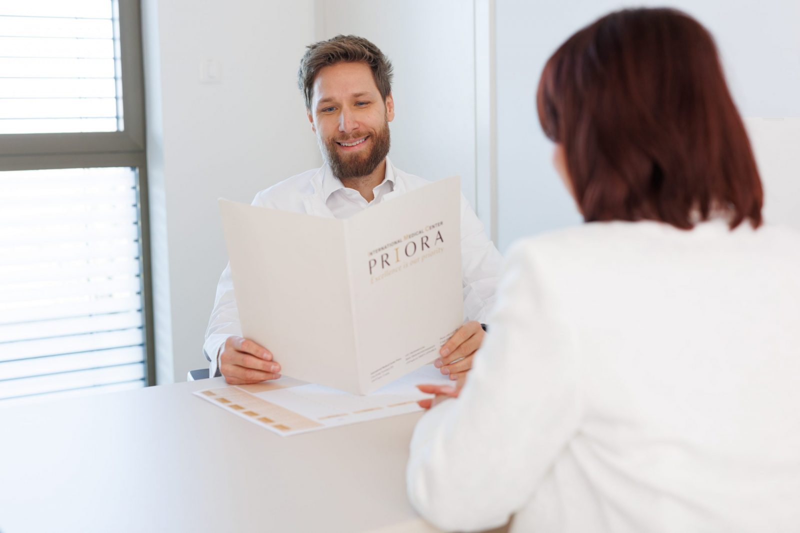 Two people in white attire at a desk; one smiles while holding a folder labeled PRIORA.